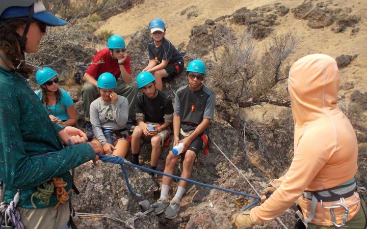 Students wearing helmets watch as an outward bound instructor provides a rock climbing lesson.